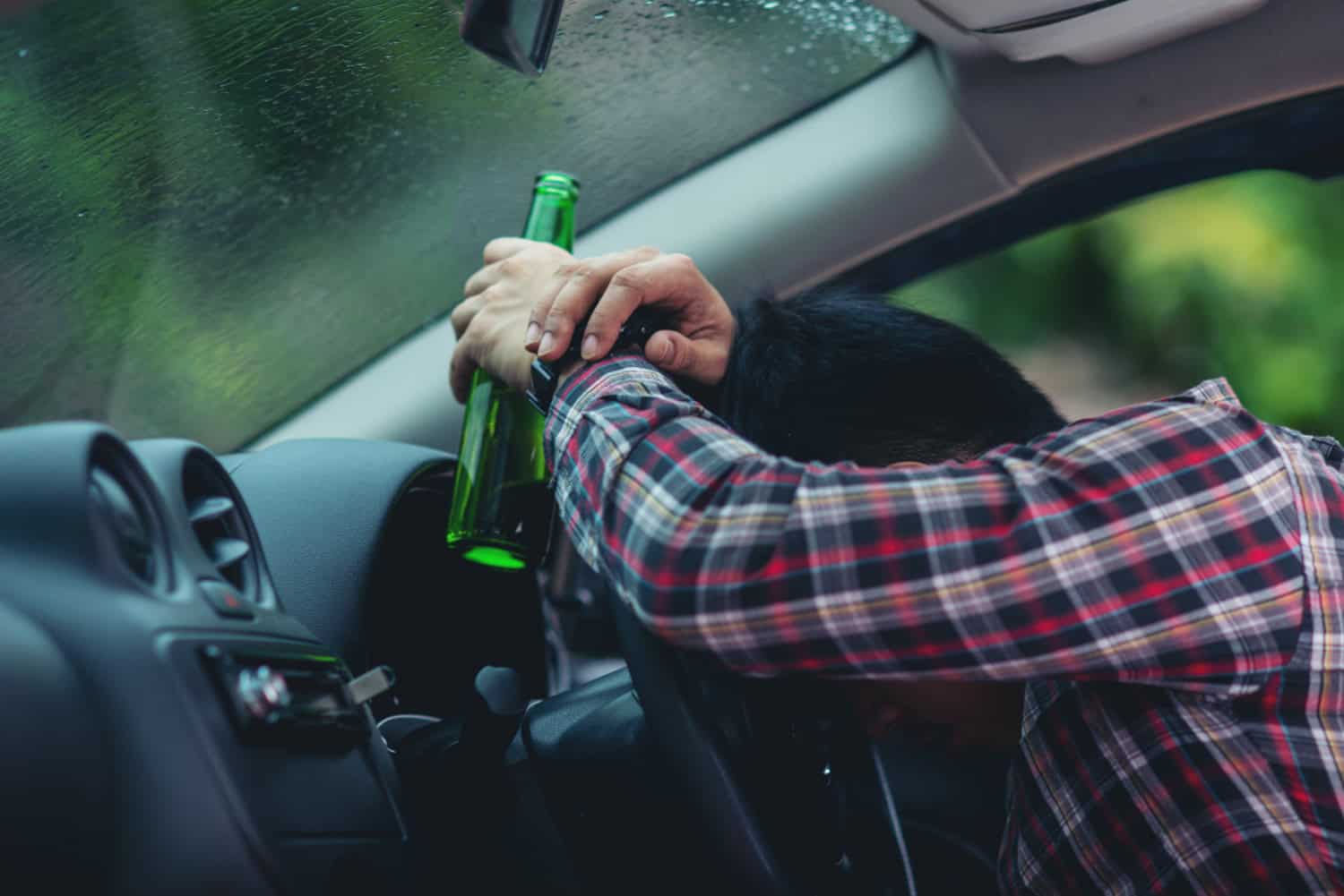 A person in a plaid shirt slumped over the steering wheel inside a car, holding an empty green bottle, suggesting intoxication.